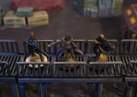 Photo of three miniatures, an assassin, a rogue trader, and a mercenary standing on a metal walkway with scifi scenery in the background.